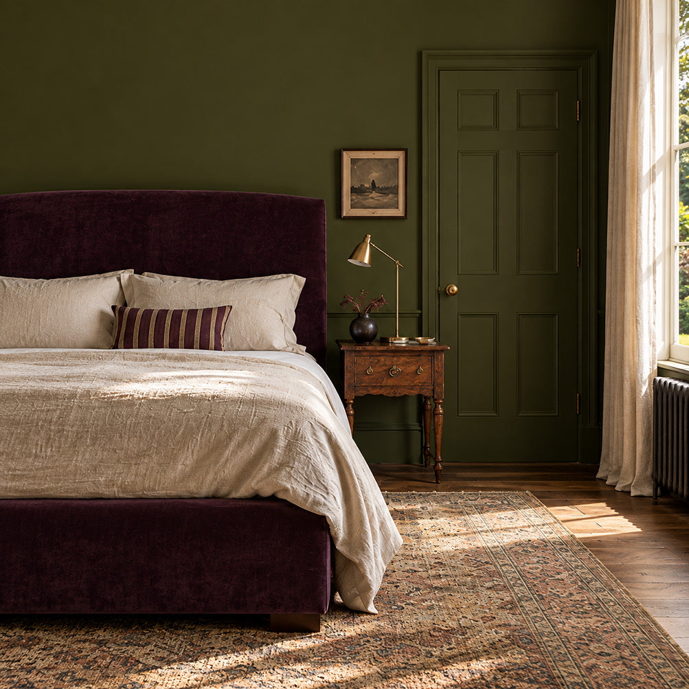 Garden Armory dark olive green bedroom walls with a plum upholstered bed, ivory bedding, and antique wood nightstand.