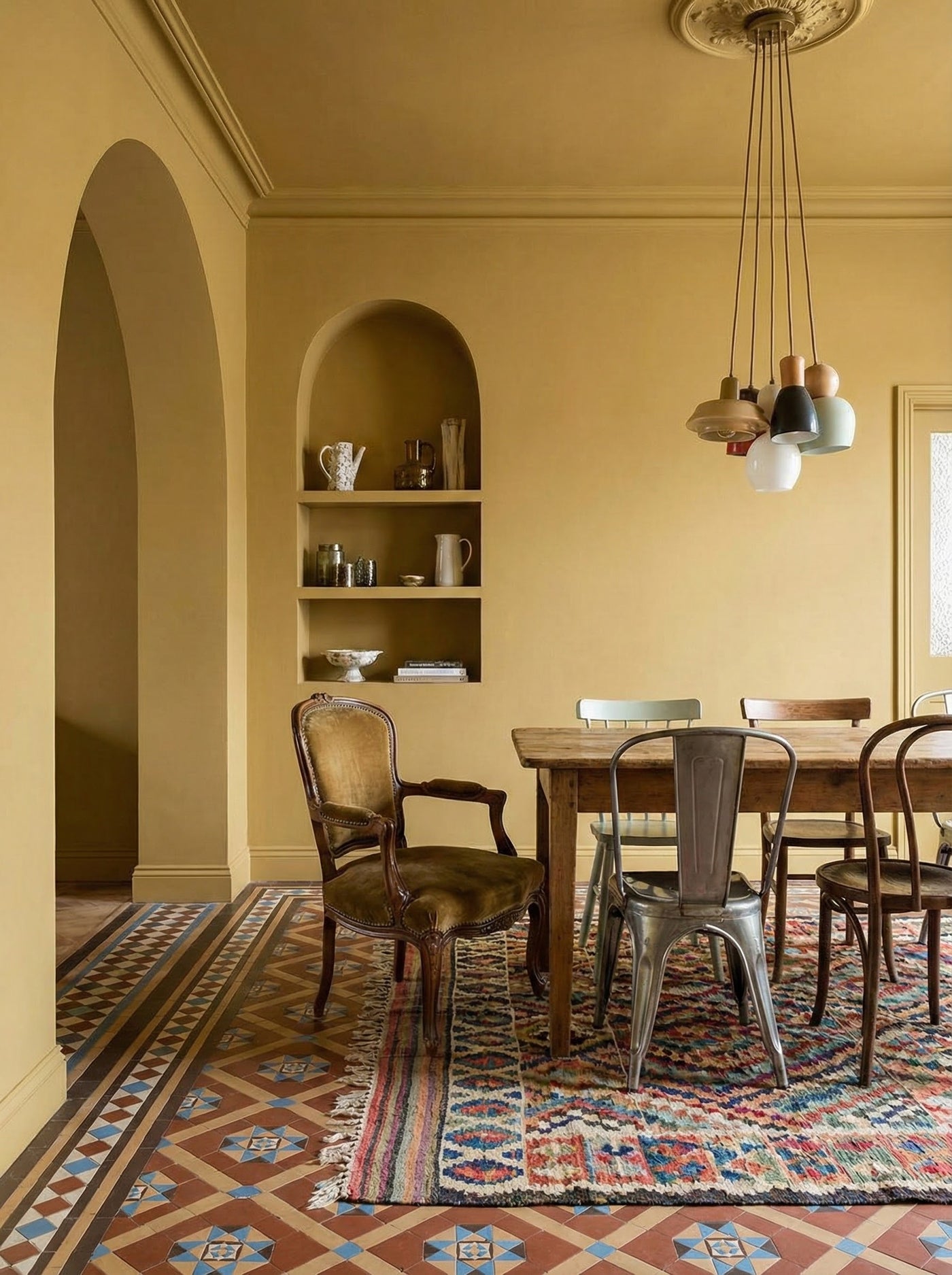 A golden yellow Dining room with a table and chairs on a patterned rug, shelves in the wall, and a pendant light.