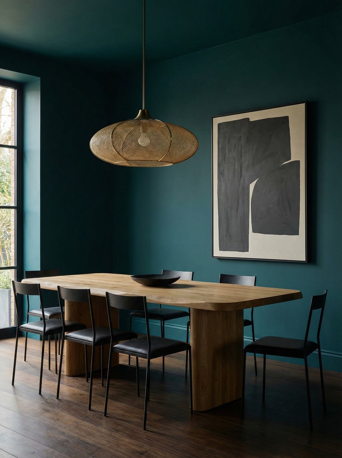 Dining room with wooden table and chairs against a moody teal painted wall with abstract art.