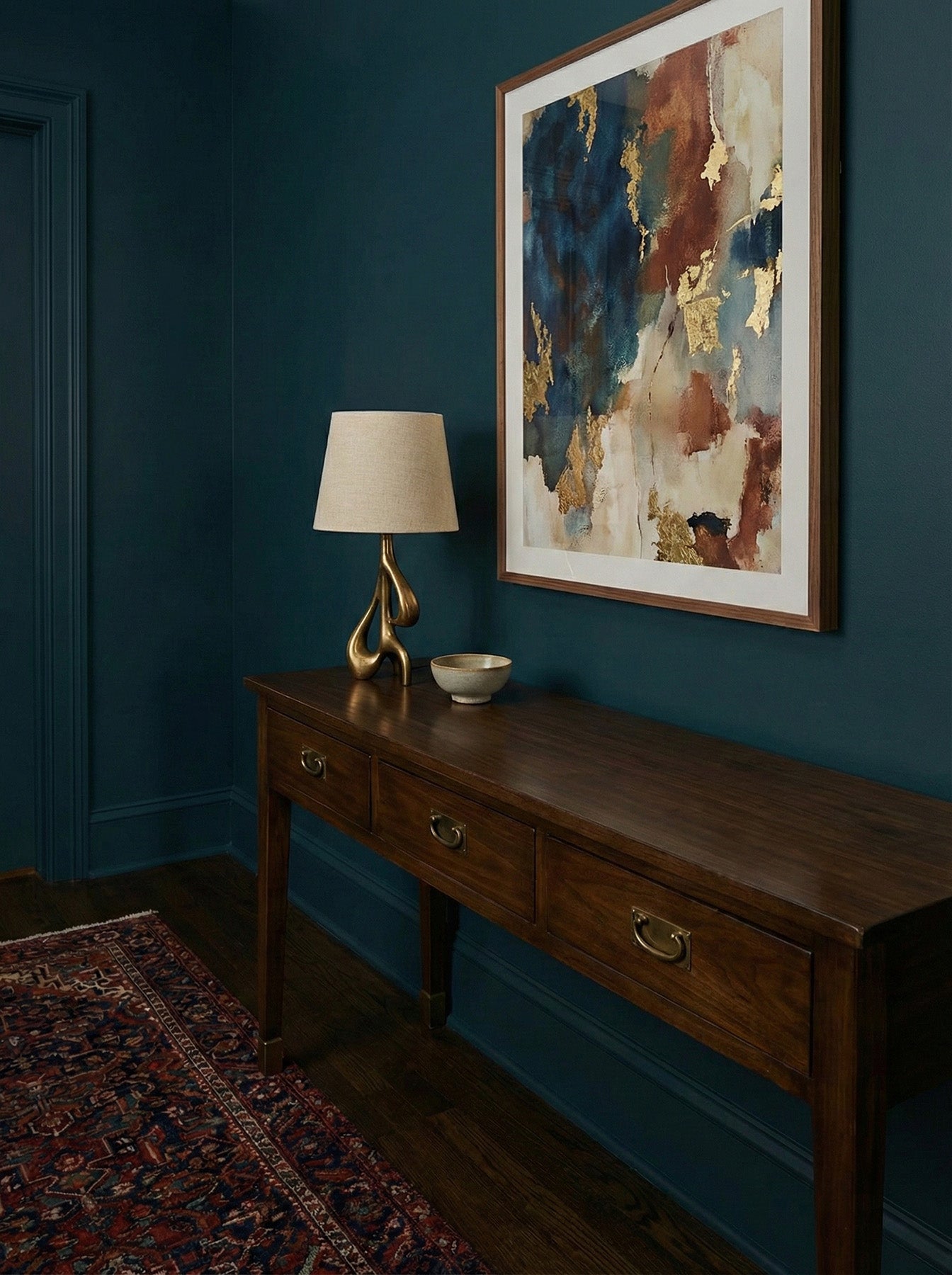 Wooden console table with lamp and decorative items against a dark blue wall with a framed abstract painting.