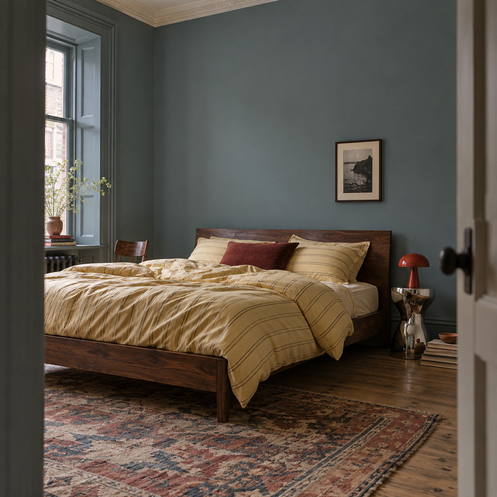 Salt Fjord muted blue-green grey bedroom walls with a walnut bed, butter-yellow bedding, and red bedside lamp.