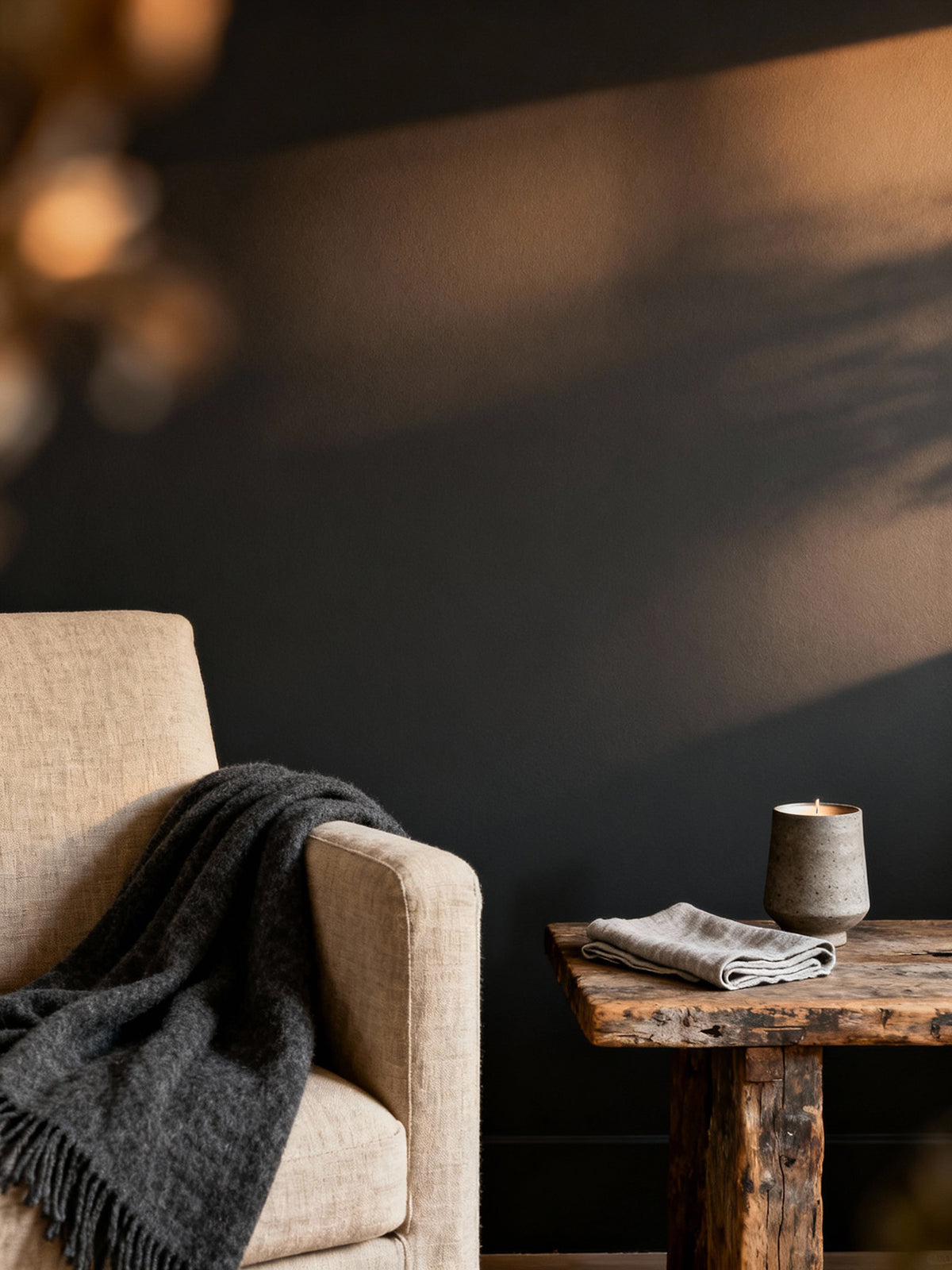 Beige armchair with a dark blanket and wooden side table with a candle and cloth against a dark wall.
